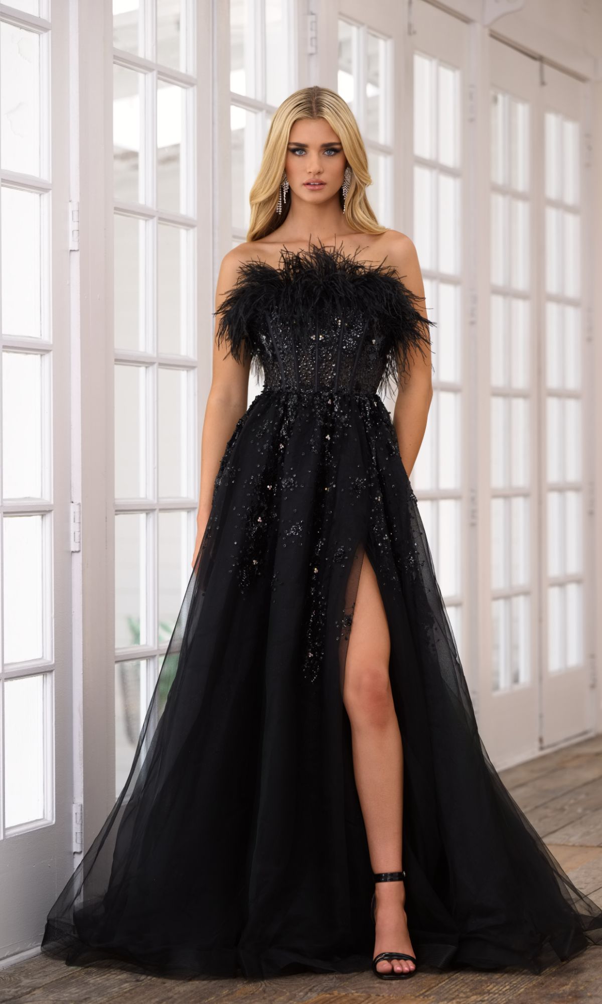 A woman in the Ava Presley 39232 Feather Long Prom Ball Gown, a black strapless dress with a high slit and feather trim, stands indoors near tall windows.